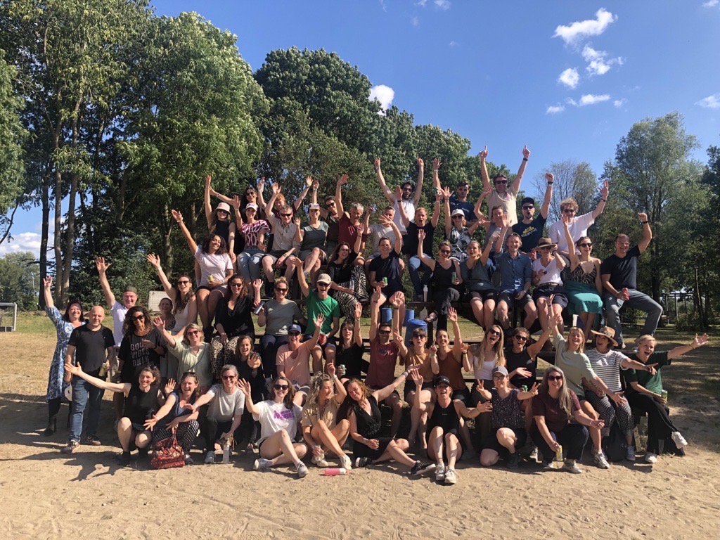 Groepsfoto van de medewerkers van Milieu Centraal in de zomer van 2025. De groep is op een strand en op de achtergrond zijn bomen te zien.